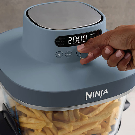 Primo piano di una persona che regola la manopola di una friggitrice ad aria Ninja azzurra, con patatine fritte visibili nel cestello in vetro.
