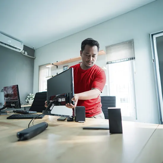 Uomo in maglietta rossa installa monitor nero su scrivania con computer e accessori in ufficio luminoso.