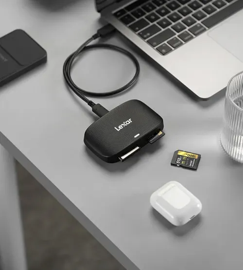 Cavo USB collega lettore Lexar a laptop su tavolo grigio. Vicino, scheda SD, custodia auricolari e bicchiere.