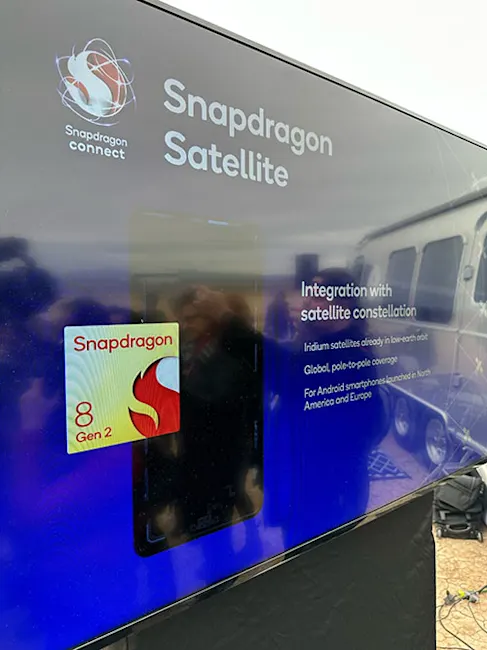 Schermo mostra "Snapdragon Satellite" con logo e scritta "8 Gen 2". Sullo sfondo, un furgone e testo su integrazione satellitare.