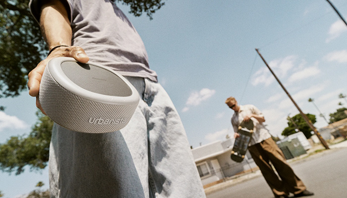 Primo piano di una persona che tiene in mano un altoparlante grigio, con una persona e uno skateboard sullo sfondo.