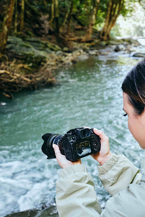 Una donna tiene una fotocamera nera di fronte a un fiume turchese e una foresta verde.