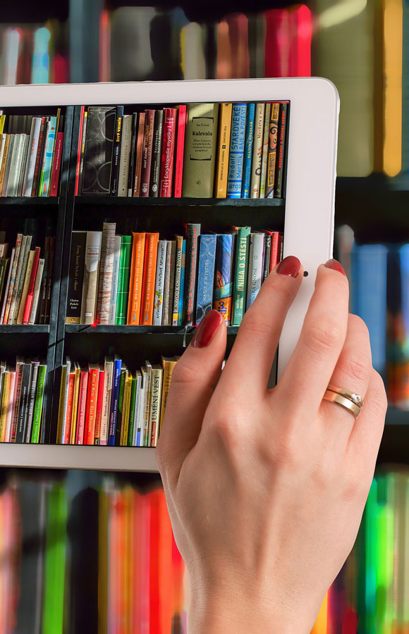 Una mano con anello tiene un tablet bianco. Sullo schermo si vedono scaffali pieni di libri colorati.