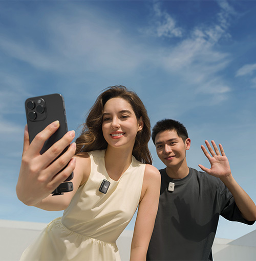 Una donna scatta un selfie con un telefono nero mentre un uomo saluta, entrambi indossano microfoni, sullo sfondo un cielo azzurro.