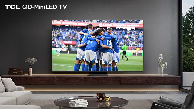 TV TCL mostra giocatori di calcio in blu che si abbracciano su campo verde. Pubblico sugli spalti. Salotto moderno.