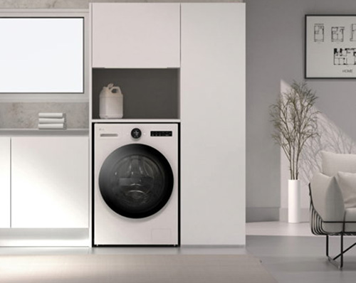 Lavatrice bianca incassata in mobili, con detersivo sopra. Dietro, pianta decorativa e sedia in stanza grigia.