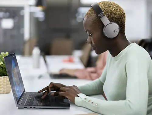 Donna con cuffie usa un laptop in ufficio. Pianta in vaso sul tavolo. Altri colleghi sullo sfondo.