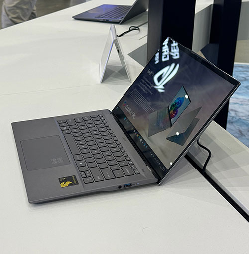 C'� un computer portatile grigio aperto su un tavolo bianco, con altri computer portatili sullo sfondo. Sullo schermo appare un logo.