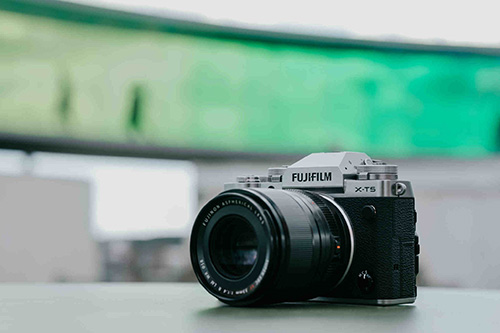 Fotocamera Fujifilm X-T5 nera e argento su una superficie, con sfondo verde e bianco sfocato.