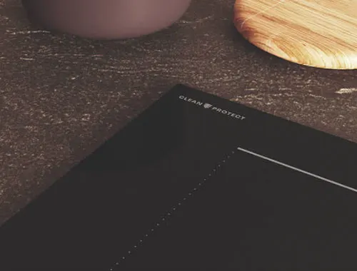 Piano cottura nero con scritta "Clean Protect", parte di ciotola marrone e tagliere di legno su piano di lavoro scuro.