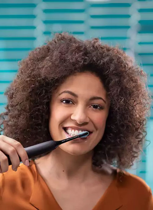 Donna sorridente si lava i denti con uno spazzolino elettrico nero. Sfondo blu orizzontale a righe. Indossa una maglietta arancione.