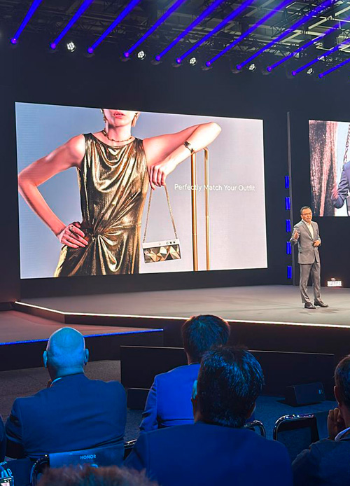 Presentazione su palco con uomo in abito e schermo con modella in abito dorato e borsa. Pubblico seduto.