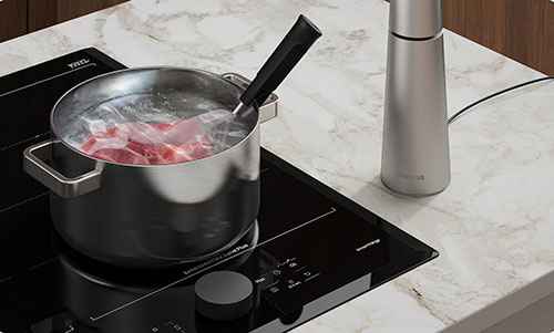 Pentola d'acciaio su piano cottura nero con acqua bollente e carne. Mestolo nero e macinino d'acciaio accanto.