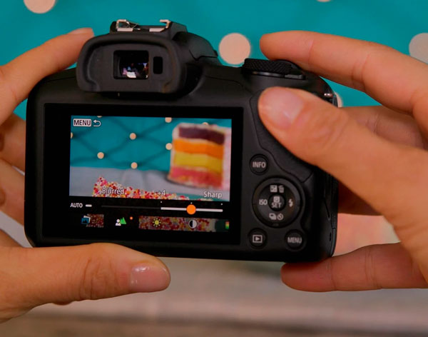 Una fotocamera nera in mano a due mani che riproduce un'immagine di una torta a strati su sfondo blu.