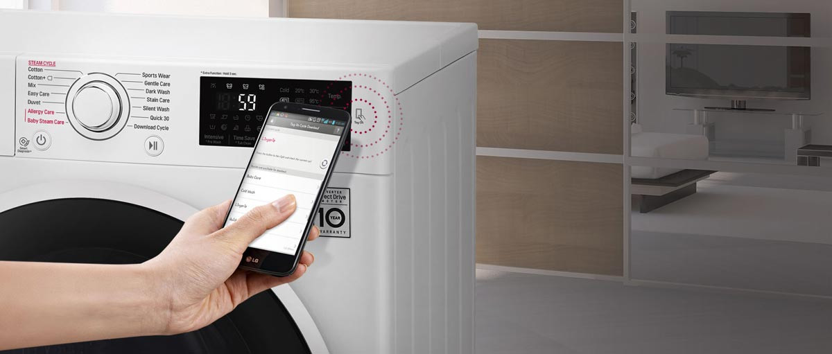 Lavatrice bianca con display digitale che mostra "59". Una mano tiene un telefono vicino al display. Stanza sfocata sullo sfondo.