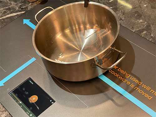 Pentola d'acciaio su piano cottura grigio con frecce azzurre. Un display nero mostra un cucchiaio arancione.