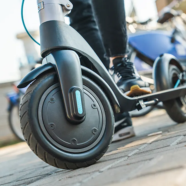 Primo piano di un monopattino elettrico grigio e nero su pavimentazione esterna. Un utente indossa scarpe da ginnastica nere.