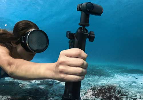Subacqueo con maschera tiene fotocamera subacquea nera sott'acqua. Il fondale marino è visibile sullo sfondo.