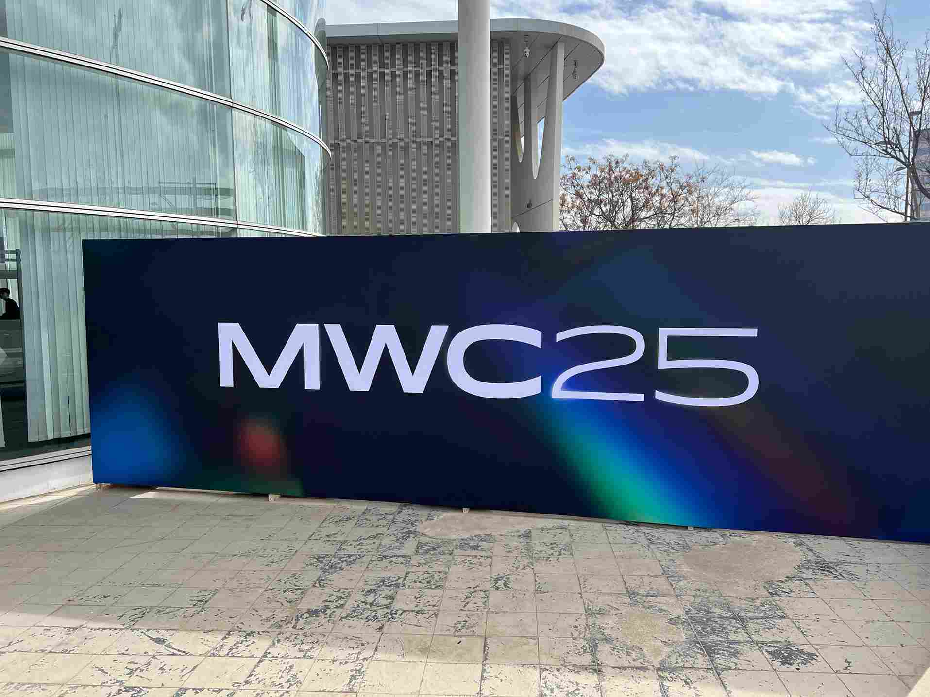 Pannello con "MWC25" in bianco su sfondo blu sfumato, esterno di edificio moderno e cielo nuvoloso.