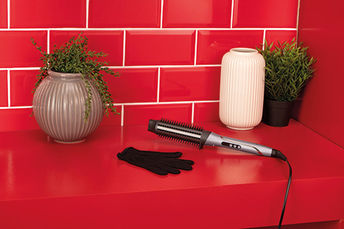 Ripiano rosso con spazzola termica, guanto nero, vaso con pianta e vaso decorativo. Sfondo con piastrelle rosse.