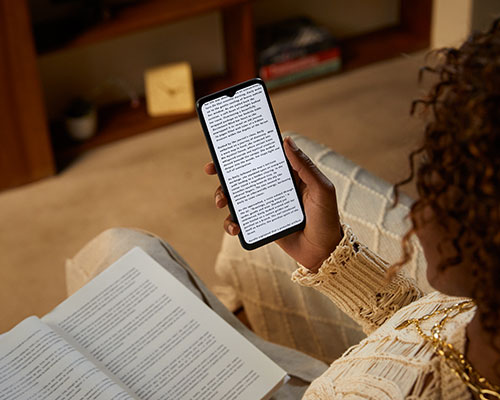 Donna seduta con libro aperto e smartphone con testo. Scaffale in legno sullo sfondo.