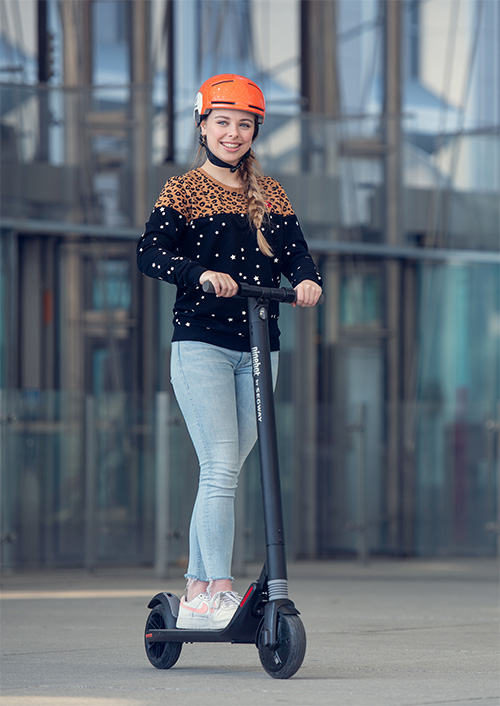 Ragazza con casco arancione su monopattino nero, indossa jeans chiari e felpa fantasia, sfondo urbano.