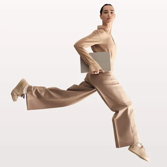 Donna in abito beige salta, tenendo una cartella. Indossa scarpe beige con lacci, su sfondo chiaro.