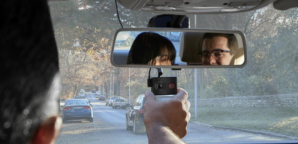 Interno di auto con specchietto retrovisore che riflette due persone. Una mano tiene una dash cam. Strada alberata sullo sfondo.