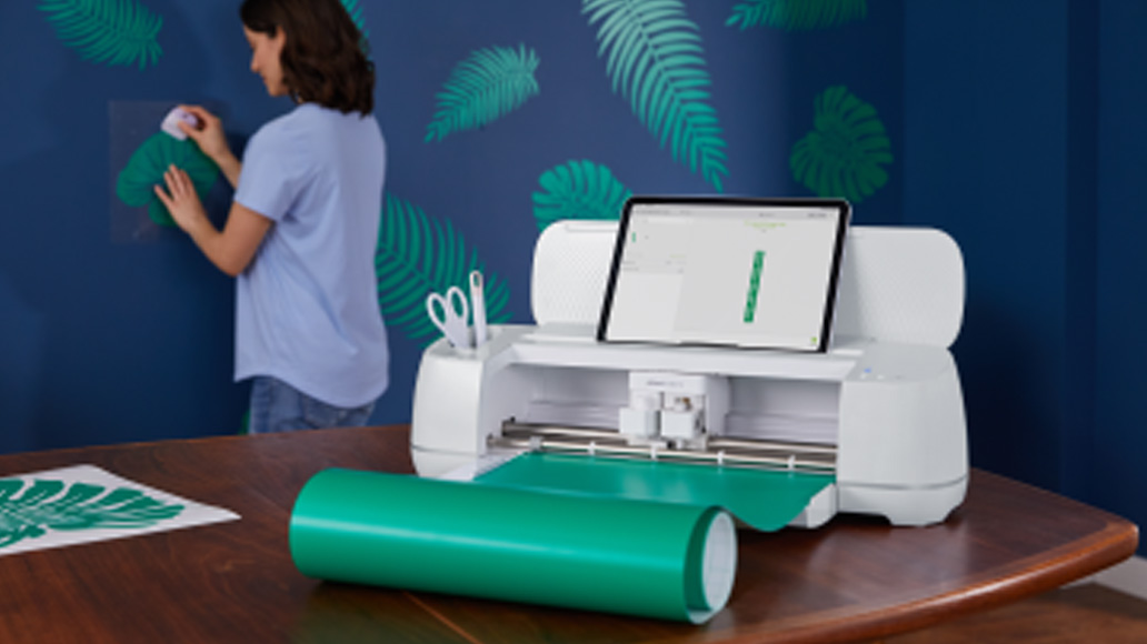 Donna applica adesivo verde su parete blu con foglie. Tavolo con plotter, tablet, vinile verde e forbici.