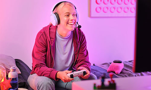 Giovane sorridente con cuffie che gioca ai videogiochi con controller. Sfondo viola con monitor e costruzioni.