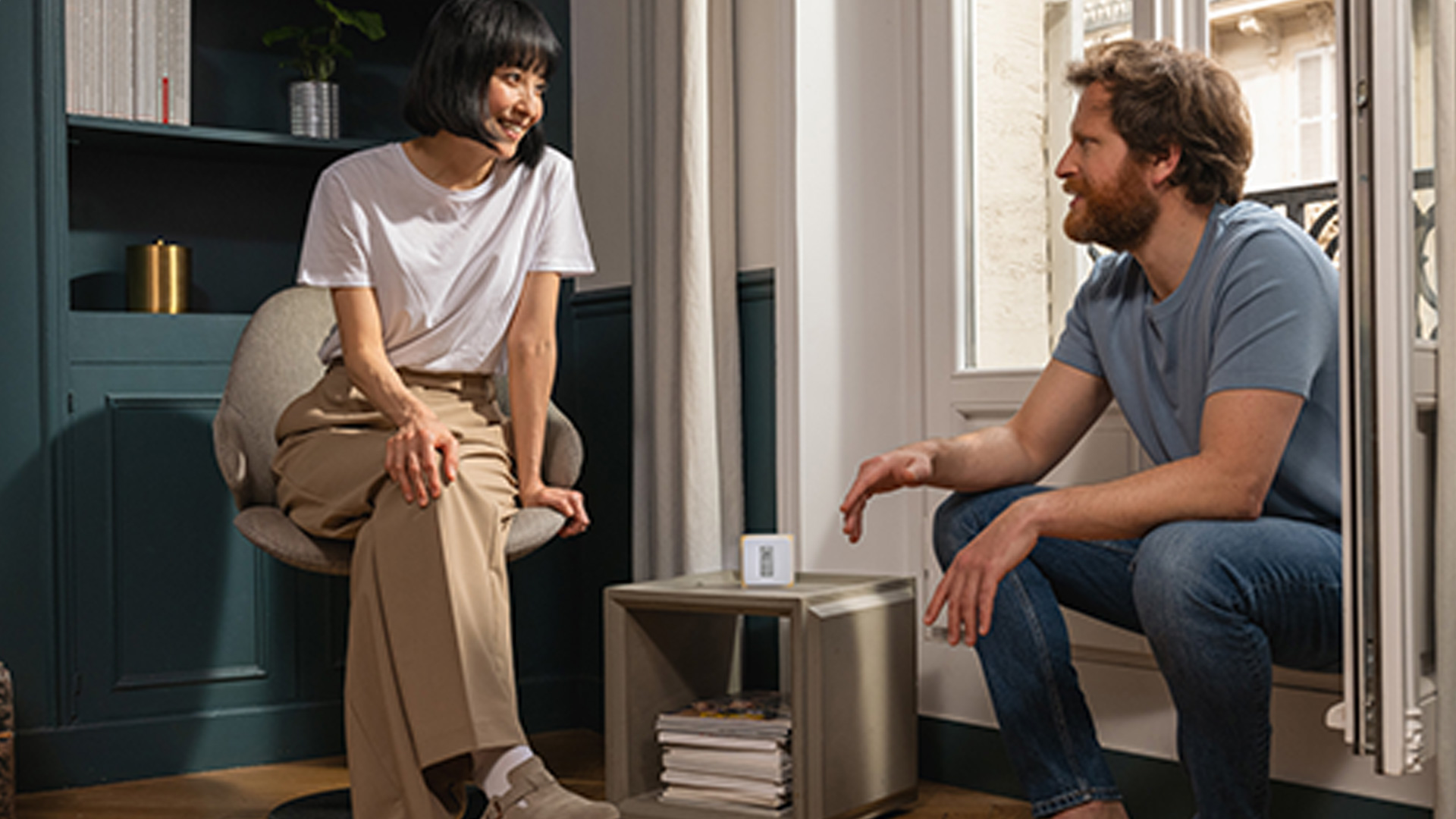 Donna seduta su una sedia beige e uomo seduto vicino alla finestra che si guardano. Tavolino con libri e un dispositivo.