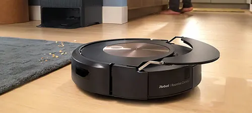 Aspirapolvere robot nero su un pavimento in legno vicino a un tappeto. Sullo sfondo si vede una base di ricarica nera.