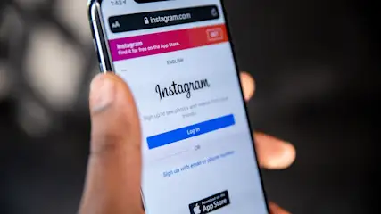 Mano che tiene un telefono con la schermata di login di Instagram su sfondo scuro. Si vedono pulsanti e testo.