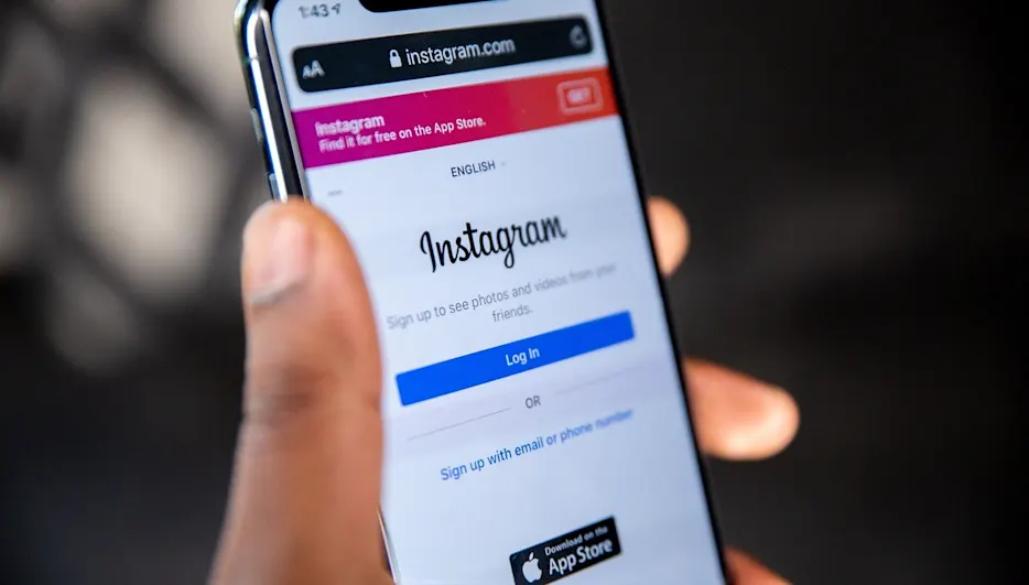 Mano che tiene un telefono con la schermata di login di Instagram su sfondo scuro. Si vedono pulsanti e testo.
