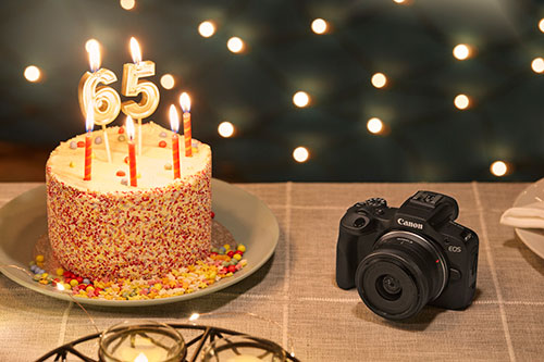 Torta con candele "65" su piatto, coriandoli. Fotocamera nera Canon a destra, luci sullo sfondo.