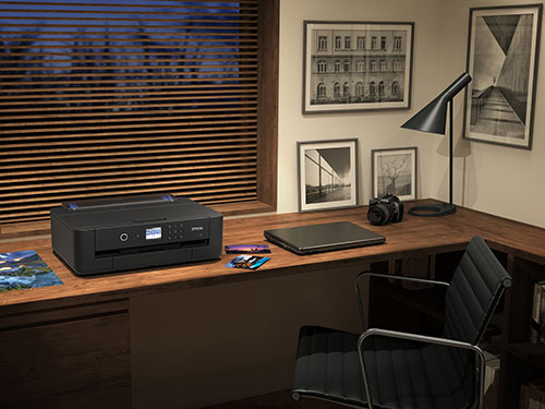 Scrivania con stampante nera vicino a una finestra con persiane, con laptop, macchina fotografica e lampada. Sul muro ci sono quadri.