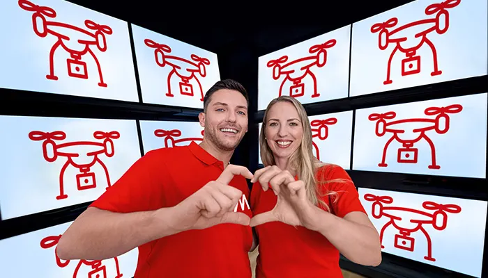 Uomo e donna sorridenti in magliette rosse formano un cuore con le mani. Schermi con icone rosse di droni sullo sfondo.