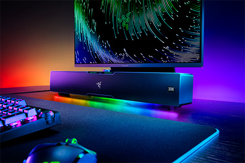 Schermo con grafica, soundbar, tastiera e mouse su una scrivania illuminata con luci colorate.