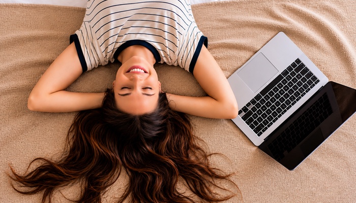 Una donna sorridente è sdraiata a pancia in su con gli occhi chiusi su una coperta beige, con un computer laptop aperto accanto a lei.