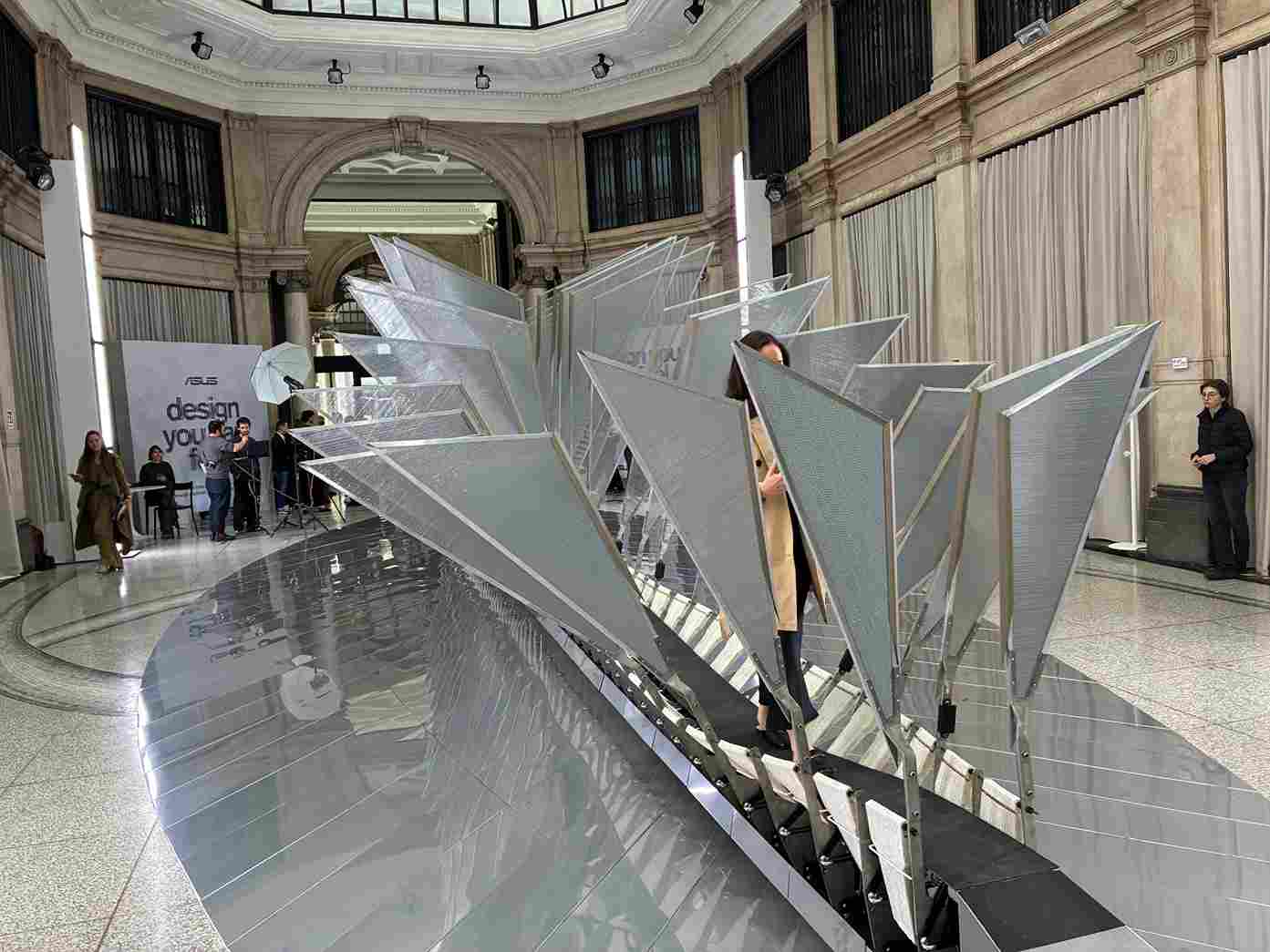 Installazione artistica geometrica argentata in spazio espositivo. Persone sullo sfondo, vetrate e logo "ASUS design you".