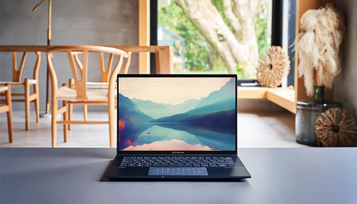 Un computer portatile su un tavolo grigio, con una finestra e sedie in legno sullo sfondo. Sullo schermo, un paesaggio montano.