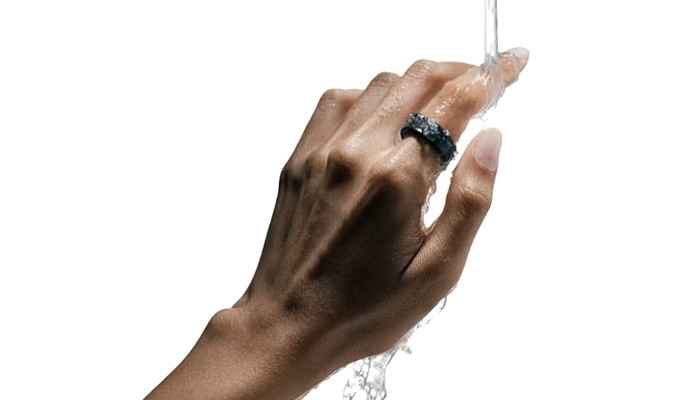 Una mano con un anello scuro è esposta all'acqua corrente su sfondo bianco.