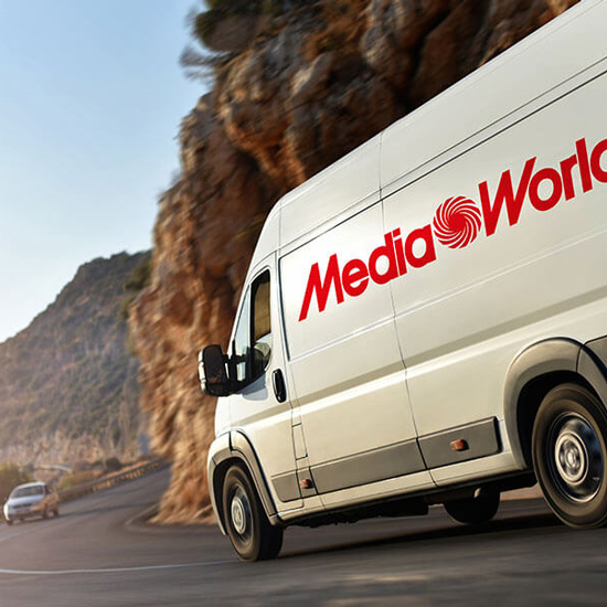 Un furgone bianco con la scritta "Media World" rossa su una strada di montagna, accanto a una roccia. Un'altra macchina è dietro.