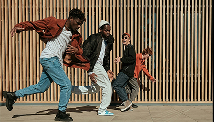 Quattro persone ballano davanti a una parete a listelli di legno. Indossano giacche, magliette e pantaloni casual.
