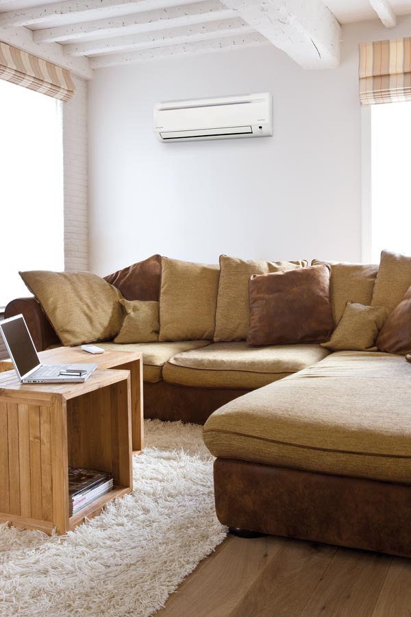 Soggiorno luminoso con divano ad angolo marrone e beige, cuscini, tavolino in legno con laptop e tappeto bianco. Condizionatore bianco a parete.