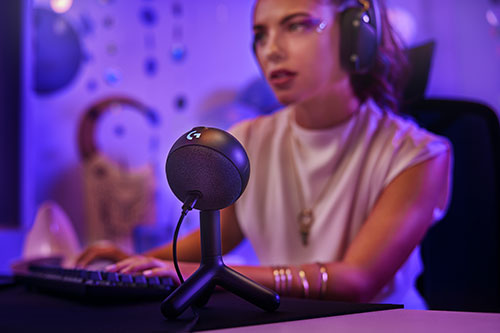 Donna con cuffie usa PC con microfono sferico. Tastiera nera in primo piano. Sfondo viola con decorazioni.