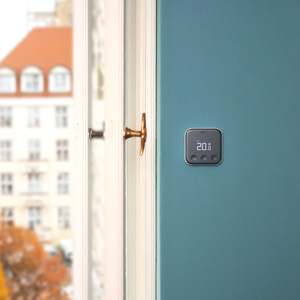 Termostato digitale grigio su parete blu, accanto a finestra bianca con vista su edificio urbano.