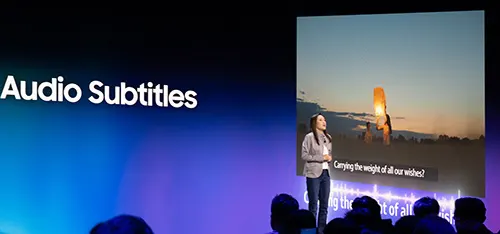 Presentazione con testo "Audio Sottotitoli" su sfondo blu sfumato. Una relatrice si trova di fronte a uno schermo che mostra un paesaggio urbano al tramonto.