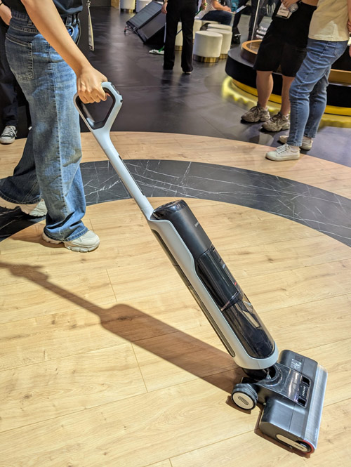 Persona usa aspirapolvere grigio su pavimento in legno. Altre persone e valigie sullo sfondo.