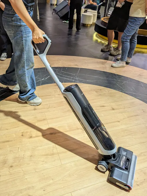 Persona usa aspirapolvere grigio su pavimento in legno. Altre persone e valigie sullo sfondo.